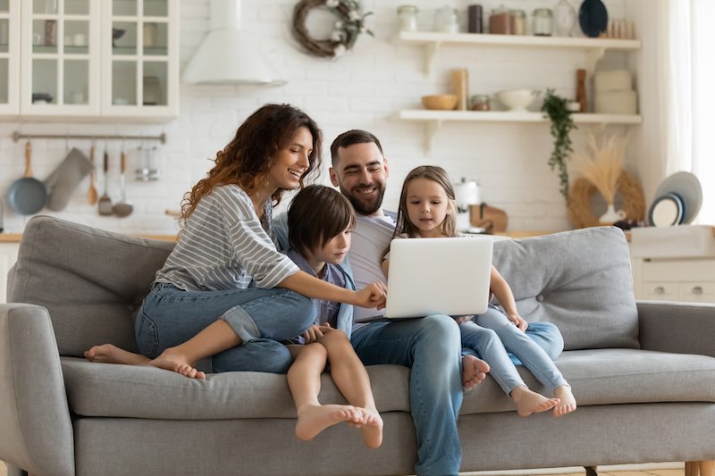 Happy young family with little kids sit on sofa in kitchen have fun using modern laptop together