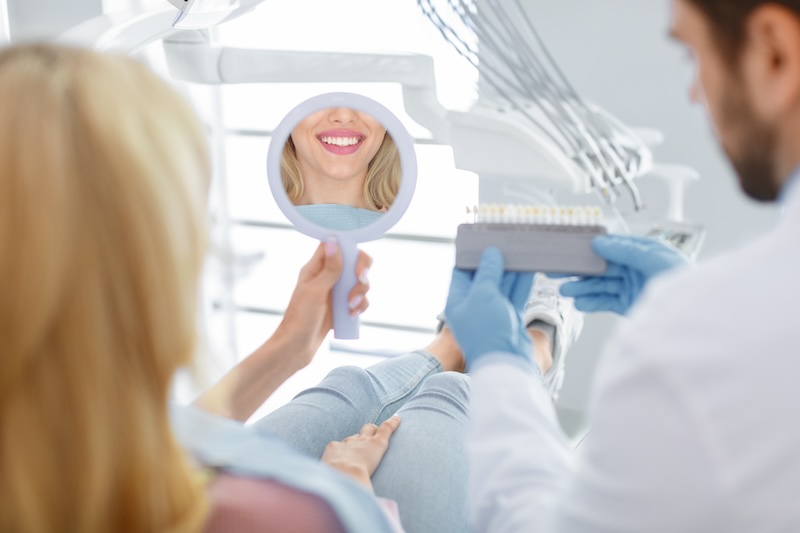 Dentist in blue medical gloves applying sample from tooth enamel scale to unrecognizable woman patient white teeth reflection in mirror to pick up right shade, teeth bleaching procedure